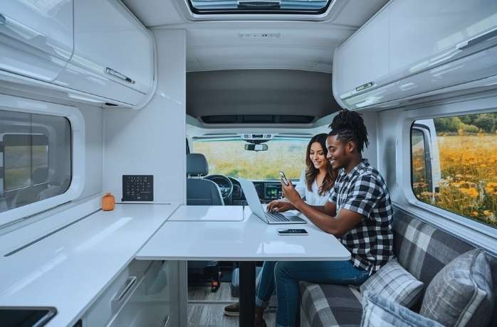 A modern RV interior with glossy white surfaces, hidden storage, and a smart home control panel. A young couple black and white race works at a fold-out desk, one editing a vlog on a laptop, the other using a smartphone to control ambient