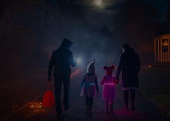 A family trick-or-treating in a suburban U.S. neighborhood at night, the kids wearing glowing LED costumes. Parents check directions on their phone while jack-o’-lanterns flicker on porches