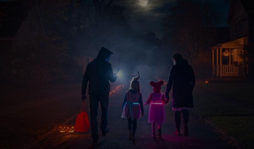 A family trick-or-treating in a suburban U.S. neighborhood at night, the kids wearing glowing LED costumes. Parents check directions on their phone